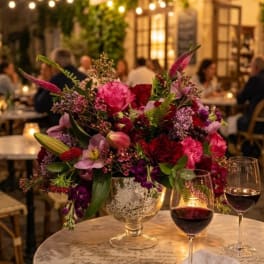 Pink and purple floral arrangement in a silver vase on a table with wine glasses