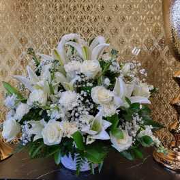 White lilies, roses, and carnations arranged in a white container with gold decor in the background.