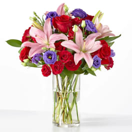 Bouquet of red roses, pink lilies, and purple blooms in a clear glass vase