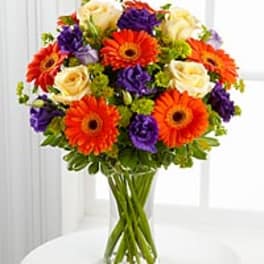 Vase arrangement of orange gerbera daisies, cream roses, and purple blooms in a clear glass vase