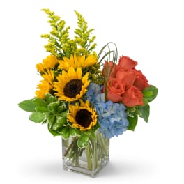 Bouquet of sunflowers, coral roses, and blue hydrangeas in a clear glass vase