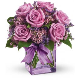 Lavender rose arrangement with small purple blooms in a square glass vase with a bow