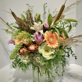 Tall rustic bouquet of peach gerbera, roses, lilies and white hydrangea in a glass vase with cattails and grasses.