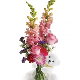 Mixed pink flower arrangement in a clear vase with a white teddy bear hugging the base
