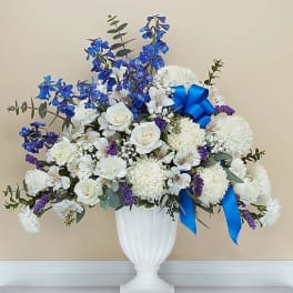 Tall white and blue sympathy flower arrangement in a white pedestal vase with a blue bow