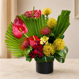 Tropical flower arrangement with red anthuriums, yellow blooms, and roses in a black vase