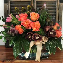 Mixed bouquet of orange roses, pink tulips, and dark orchids in a glass vase