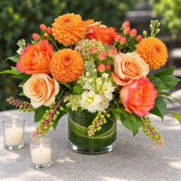 Orange roses and dahlias arranged in a glass vase with candles nearby