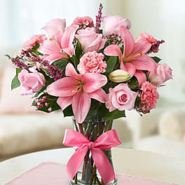 Pink lilies and roses arranged in a glass vase with a pink ribbon