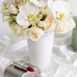 White roses and orchids arranged in a white vase with a red lipstick in front