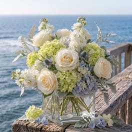 White roses and pale hydrangeas in a clear glass vase by the sea