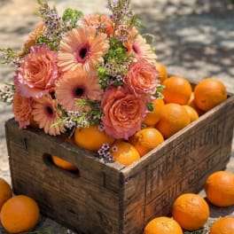 Peach and pink flower bouquet arranged in a wooden crate with oranges