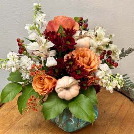 Autumn floral arrangement with orange roses, burgundy mums, white blooms, and small pumpkins in a glass vase