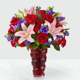 Bouquet of red roses, pink lilies, and purple flowers in a red glass vase