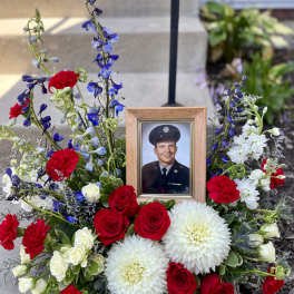 Memorial floral arrangement with a framed portrait, red roses, white blooms, and blue flowers