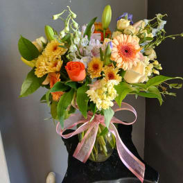 Mixed bouquet of peach, yellow, and white flowers in a glass vase with pink ribbon