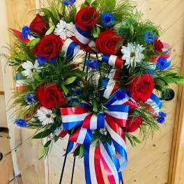 Red roses and white daisies with blue accents on a ribboned standing wreath