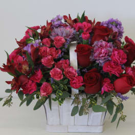 Bright basket arrangement of red roses, pink carnations, and purple blooms in a white wooden handled container