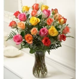 Tall glass vase filled with assorted pink, yellow, orange, and red roses with white filler flowers.