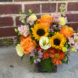 Mixed bouquet with sunflowers, orange roses, and pastel blooms in a glass vase