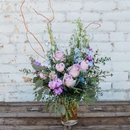Lavender roses and purple flowers arranged in a clear glass vase.