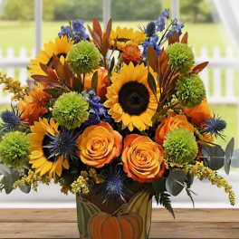 Autumn bouquet with sunflowers, orange roses, green mums, and blue accents in a pumpkin-decorated vase.