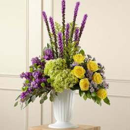Tall floral arrangement with yellow roses, purple blooms, and green hydrangeas in a white vase