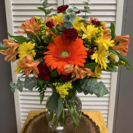Mixed bouquet with orange gerbera daisies, yellow daisies, and red carnations in a glass vase