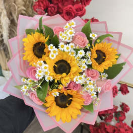 Hand-tied bouquet of yellow sunflowers, small white daisies, and pink roses wrapped in layered pink paper.