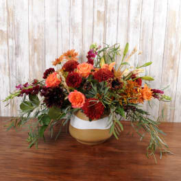 Low arrangement of orange and burgundy flowers with trailing greens in a round ceramic vase