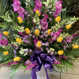Large mixed floral arrangement with yellow roses, pink gladiolus, and purple daisies in a white vase