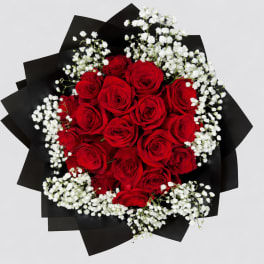 Round bouquet of red roses and white filler flowers wrapped in black paper.