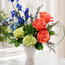 Bouquet of coral roses, blue flowers, and pale green hydrangeas in a white vase