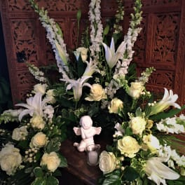 White floral funeral arrangement with lilies and roses around a small angel figurine