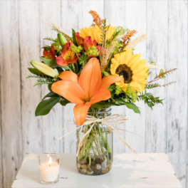 Orange lilies and sunflowers in a glass vase with a candle nearby.