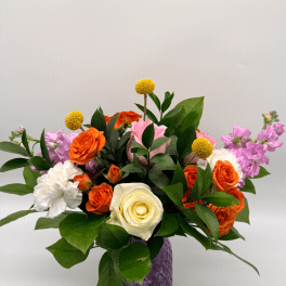 Colorful arrangement of orange roses, white rose and carnation, and pink blooms in a purple glass vase
