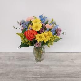 Mixed bouquet of yellow daisies, orange rose, and pastel flowers in a clear glass vase
