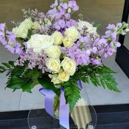Vase arrangement of cream roses and light purple flowers with ferns and a lavender ribbon