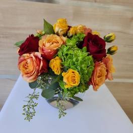 Low glass vase of orange, yellow, and red roses with bright green hydrangea