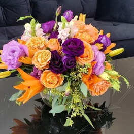 Low arrangement of orange lilies and roses with purple blooms in a clear glass bowl on a black table