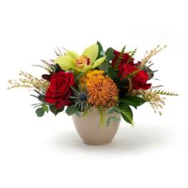Low arrangement of red roses, green cymbidium orchid, and orange protea in a beige bowl vase