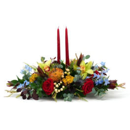 Low centerpiece with red roses, yellow pincushion blooms, green orchids and blue flowers around two tall red candles
