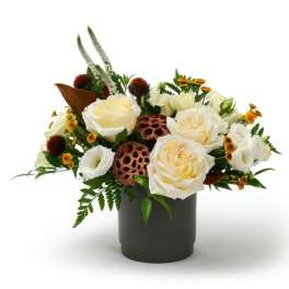 Low arrangement of cream roses, white blooms, and yellow accents in a dark cylinder vase with lotus pods.