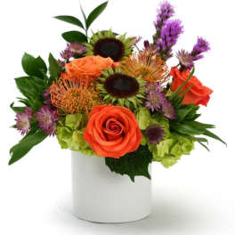 Compact arrangement of orange roses, green hydrangeas, and purple blooms in a white cylinder vase