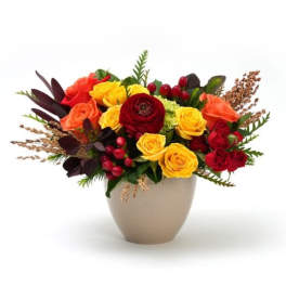 Low arrangement of yellow, orange, and red roses with berries in a beige vase