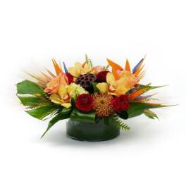 Low tropical arrangement of orange, yellow, and red flowers in a round glass vase