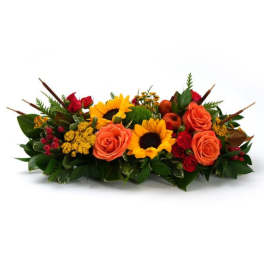 Low centerpiece of orange roses, yellow sunflowers, and red accent blooms in an elongated design
