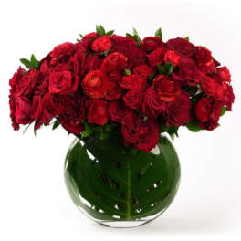 Low round arrangement of red roses and ranunculus in a clear glass vase lined with a large green leaf.