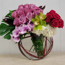 Low glass vase arrangement with lavender roses, red roses, green orchids, white hydrangeas, and purple orchids.