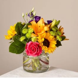 Mixed bouquet in a clear glass vase with a pink rose, sunflower, lily, and purple blooms
